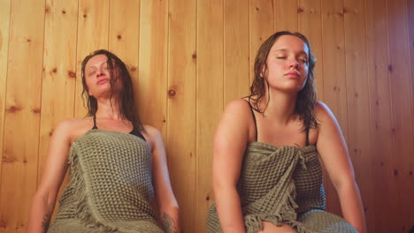 two friends wrapped in textured towels sit against wooden panel walls inside sauna, cooling off after steam session, damp hair resting on shoulders and eyes closed in calm relief