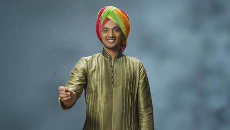 young happy indian man in turban and traditional clothes smiling at camera and holding incense stick