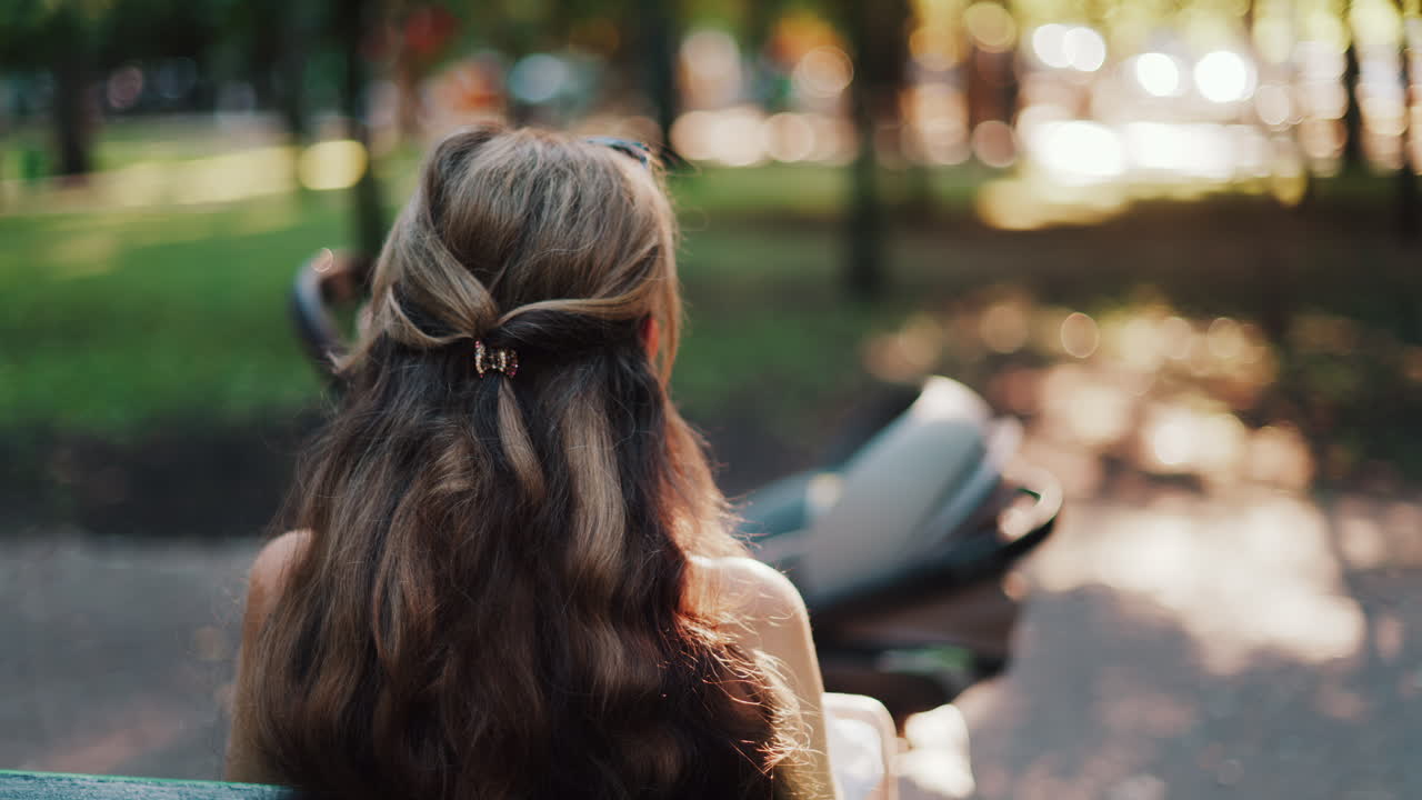 A mother with long wavy hair siting on a bench, breastfeeding her baby in a peaceful park, next to a modern baby stroller
