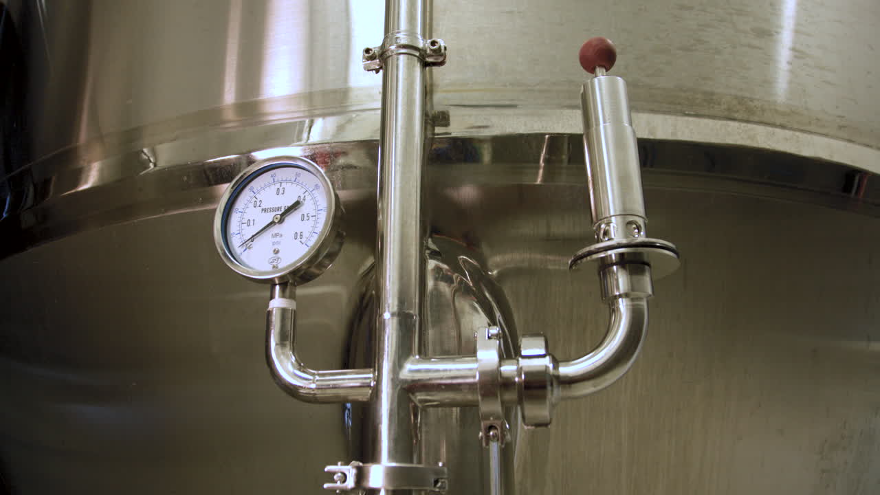 Rack focus from a close-up of a pressure gauge and pipe mounted on a stainless steel fermentation tank in a brewery to a blurry view