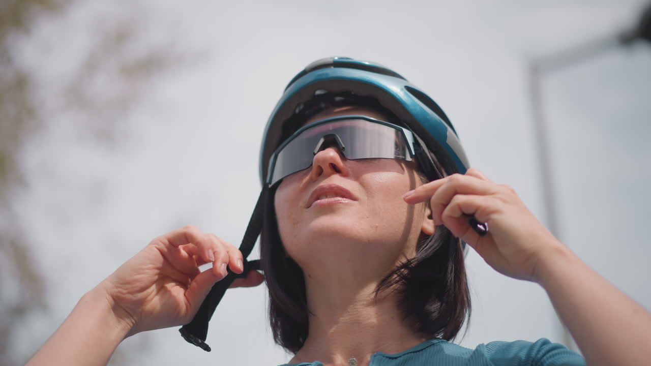 woman adjusting helmet and sunglasses on treelined street, focused expression, modern cycling gear with reflective visor, tightening chin strap, poised to start commute or training ride