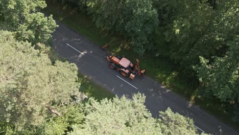 an orange tractor equipped with maintenance attachments is parked beside a forest road. the vehicle is likely performing roadside vegetation control, with green trees surrounding the asphalt road.