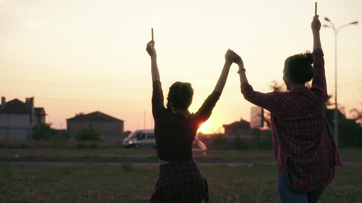 Back View Of Two Girlfriends Walking Forward And Waving With Firework Candles During Sunset