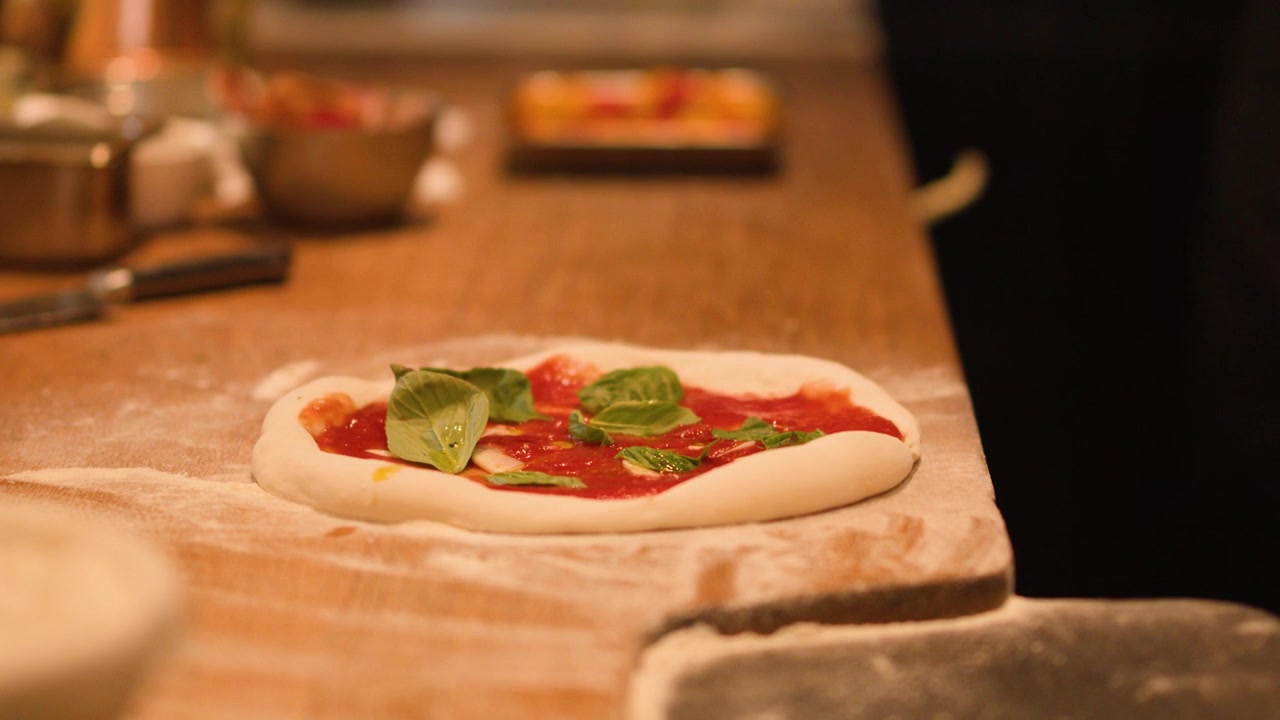 Primo piano di impasto crudo per pizza con salsa di pomodoro e basilico su un bancone di legno.