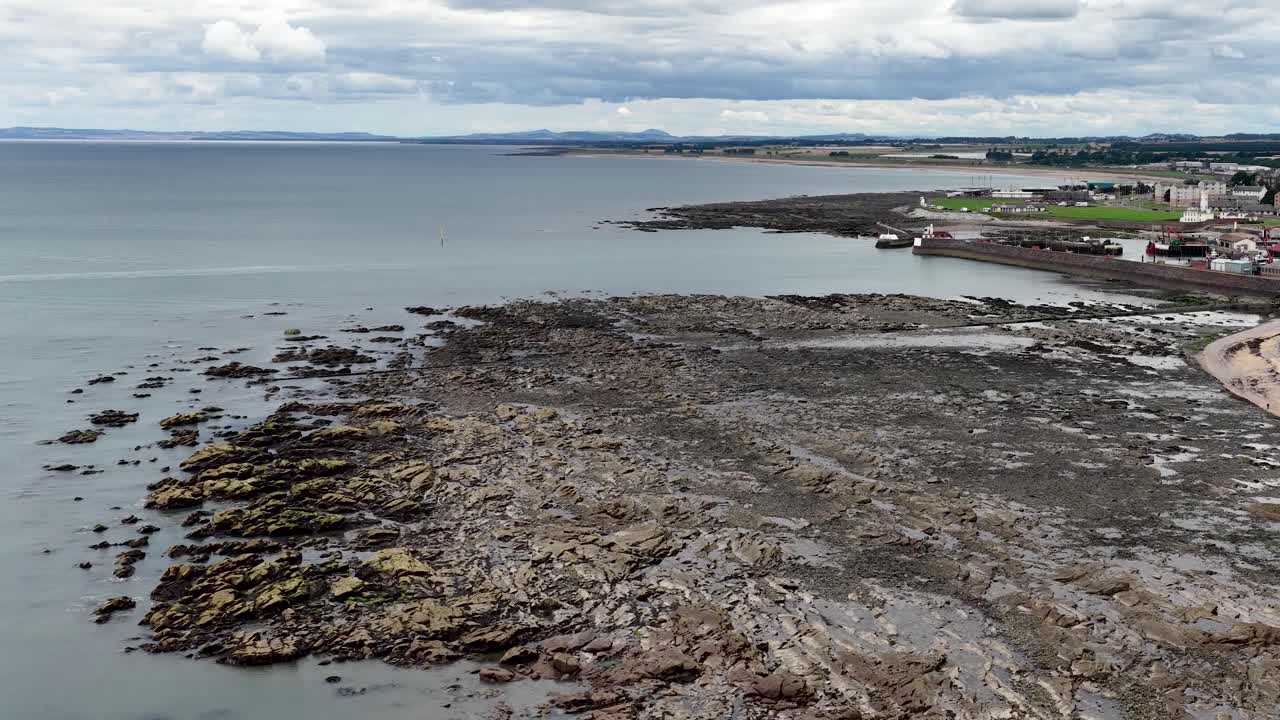 Drone camera slowly pans across rocky shoreline, coastal town, and North Sea under cloudy daylight
