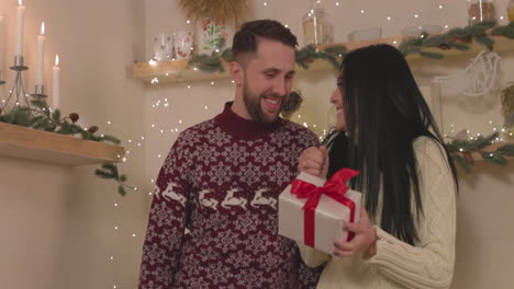 Happy-Woman-Holding-A-Christmas-Gift-Box-And-Kissing-Her-Boyfriend-At-Home-1