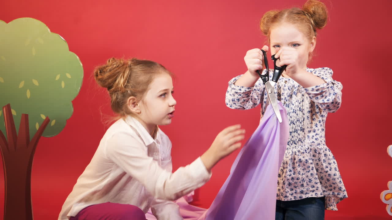 two little girls are cutting fabric of lilac colour by scissors on the background of the red walls of the studio with scenery