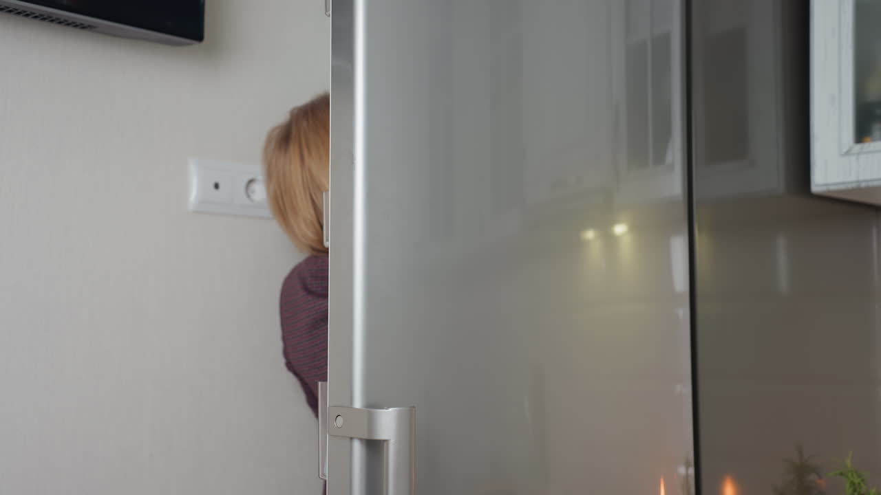 mujer abre el refrigerador para recuperar el artículo, la puerta elegante refleja el fondo de la cocina con iluminación cálida, enchufe de pared visible cerca del electrodoméstico, diseño interior moderno con paredes de azulejos
