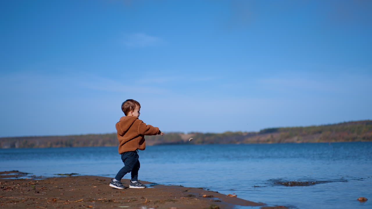 Little toddler boy approaching the river. Caucasian kid throws two stones into water.