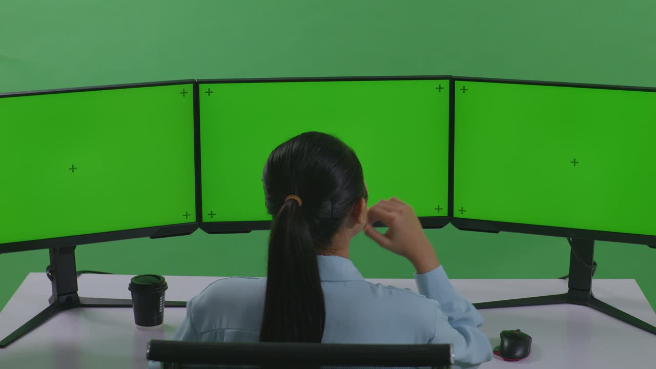 녹색 스크린 스튜디오에서 모의 여러 컴퓨터 모니터와 함께 작업하는 동안 무언가에 대해 생각하는 아시아 여성의 뒷면