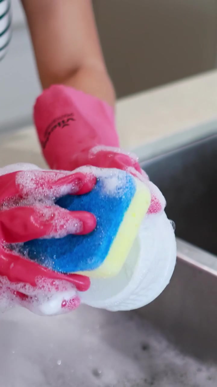 Hands in pink gloves wash a dish with a sponge in a kitchen sink under bright lighting