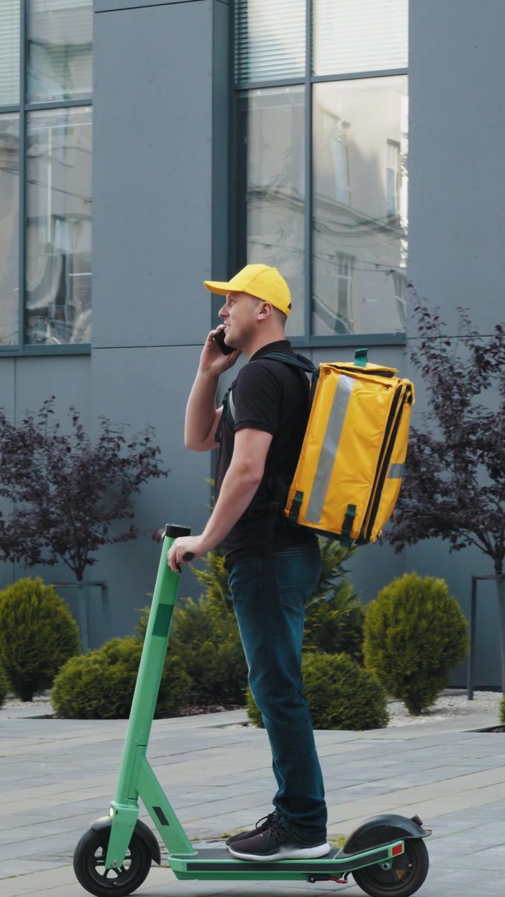 Ein Lieferant mit gelber Kappe und Rucksack telefoniert, während er einen Elektroroller fährt.
