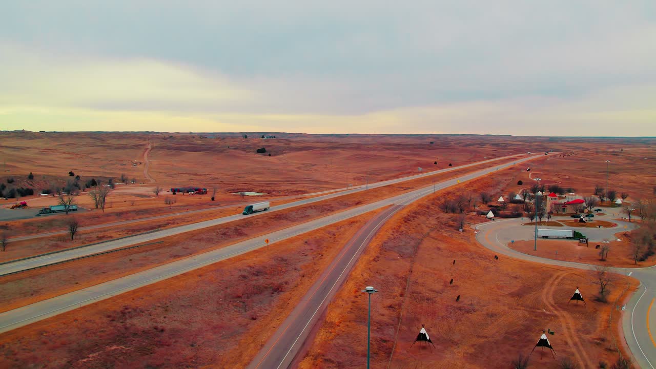 Wide aerial reveals a semi truck cruising along scenic Colorado highways near teepees and a desert-like rest area on rolling red plains.