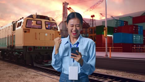 asian female with her smarphone with freight cargo train at port, she raises her fist up with screaming goal