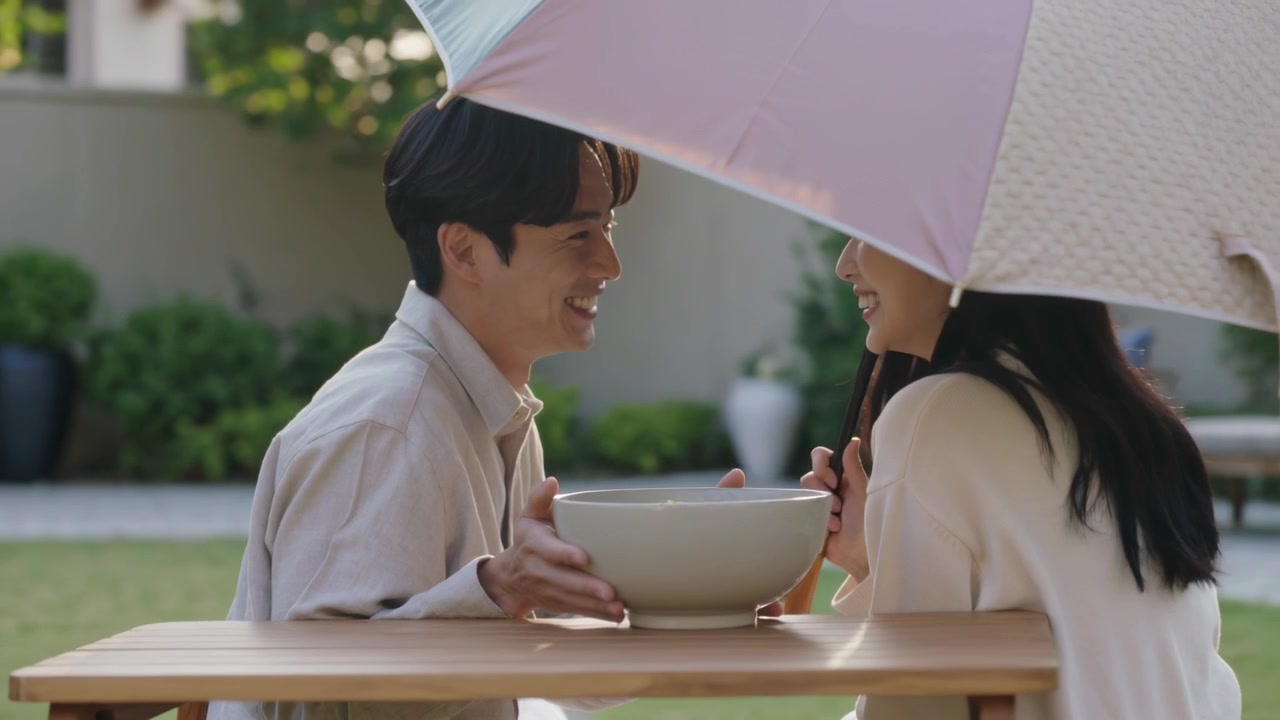 穿着浅色衬衫的男人伸手把碗递给女人，在撑着伞的后院桌旁分享。