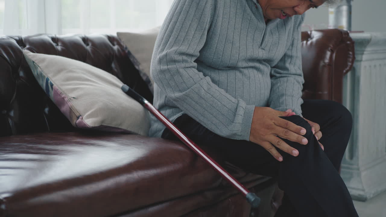 심각한 다리와 무릎 통증으로 고통스러운 표현을 하는 노인은 일어나서 소파에 앉아 집안을 돌아다니는 데 어려움을 겪습니다.