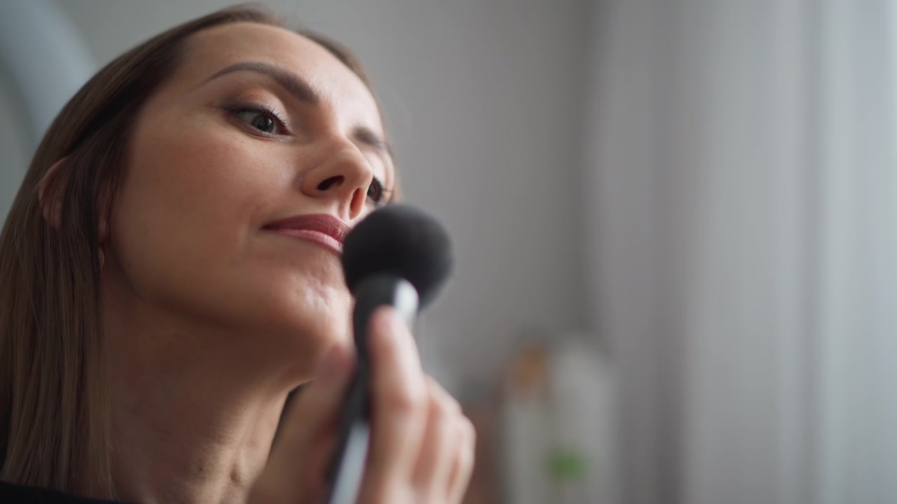 Close up of woman applying makeup with soft brush in bathroom setting. Woman holds and moves black powder brush near her face neck visible as woman focuses on grooming routine. Background