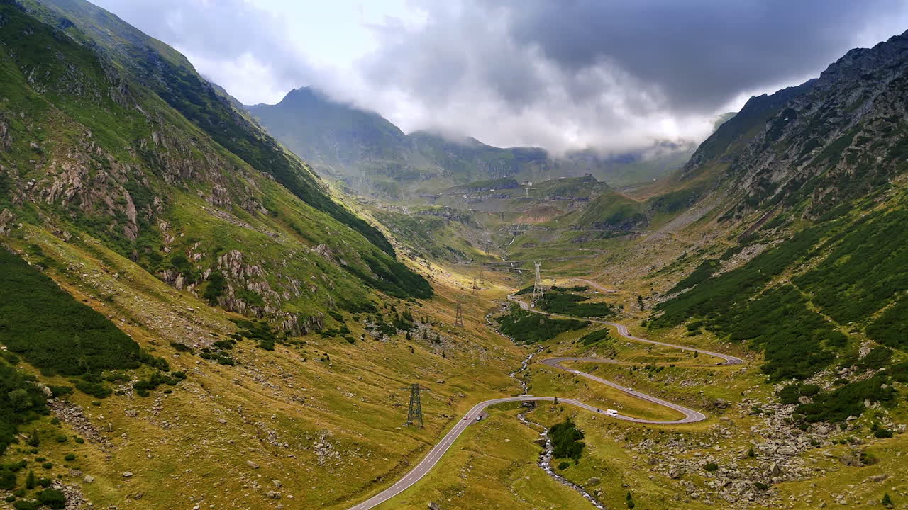 Cars move by the wavy road among the verdant mountains. Aerial view on the famous Transfagarash highway in Romania. Grey cloudscape in the sky