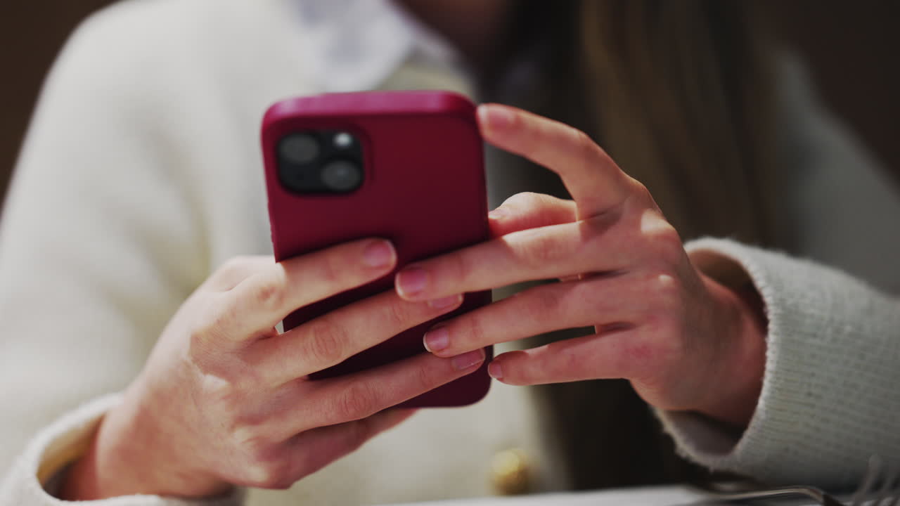 Close up of a woman's hands holding and using a smartphone with a burgundy case at a restaurant