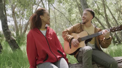 Glückliches-Afroamerikanisches-Paar,-Das-Auf-Einem-Baumstamm-Sitzt-Und-Im-Wald-Gitarre-Spielt,-Zeitlupe