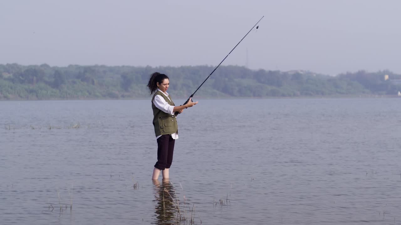 Seorang gadis Asia Selatan mengenakan rompi luar ruangan berdiri memancing di air danau yang dangkal, dengan hati-hati menggulung tali pancingnya di siang hari yang tenang