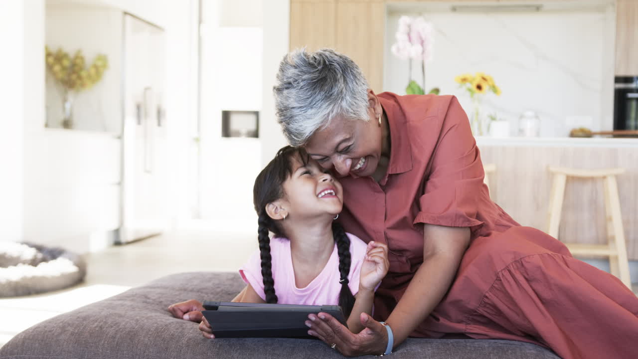Biracial grandmother laughs with a biracial granddaughter holding a tablet, with copy space