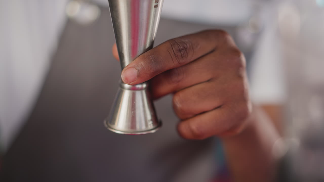 Barman pours juice into jigger in bar closeup. Black bartender measures ingredient for refreshing cocktail in club. Barkeeper makes exotic drink