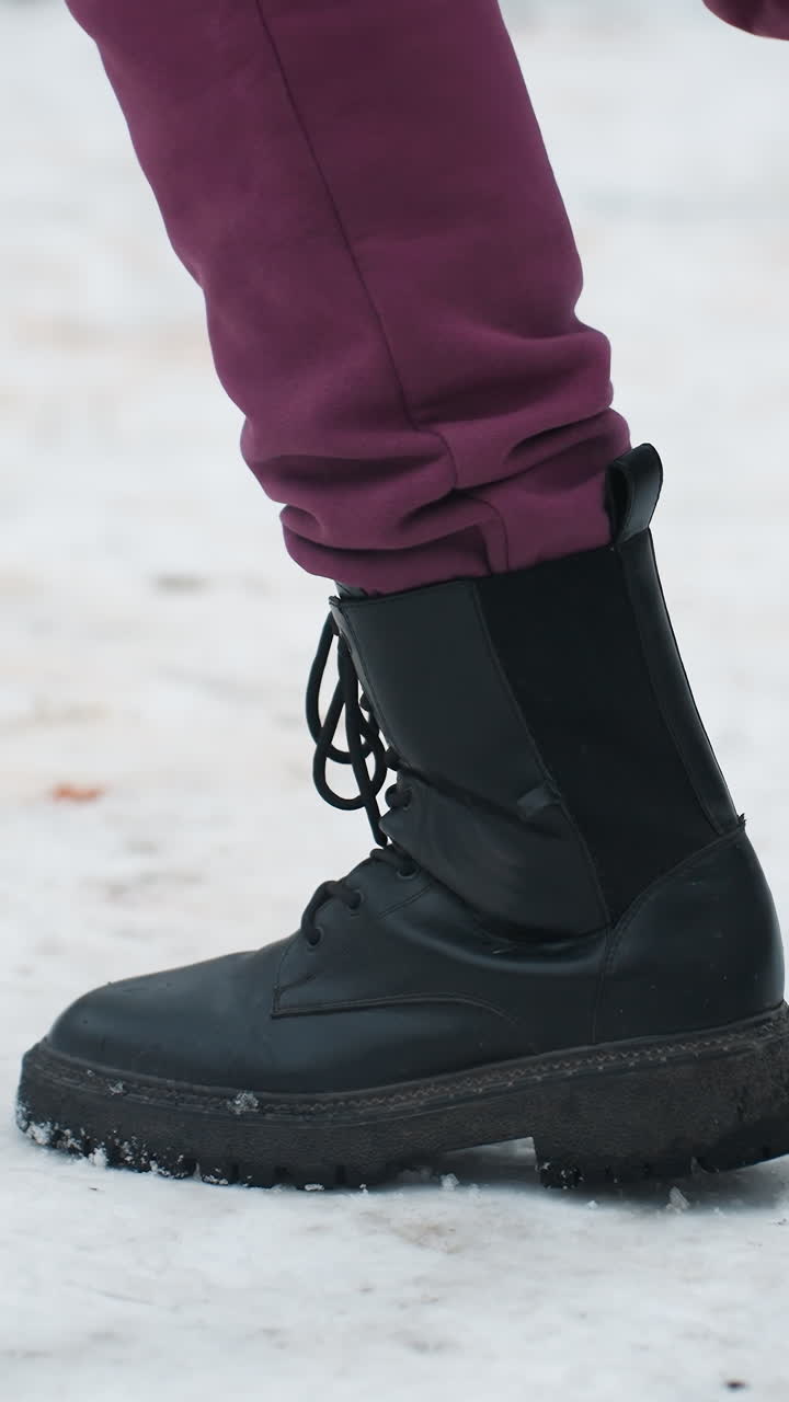 vista lateral de un individuo con botas negras saltando juguetonamente en el suelo cubierto de nieve con follaje seco esparcido, el entorno de invierno captura el movimiento, el calzado y la textura del terreno helado