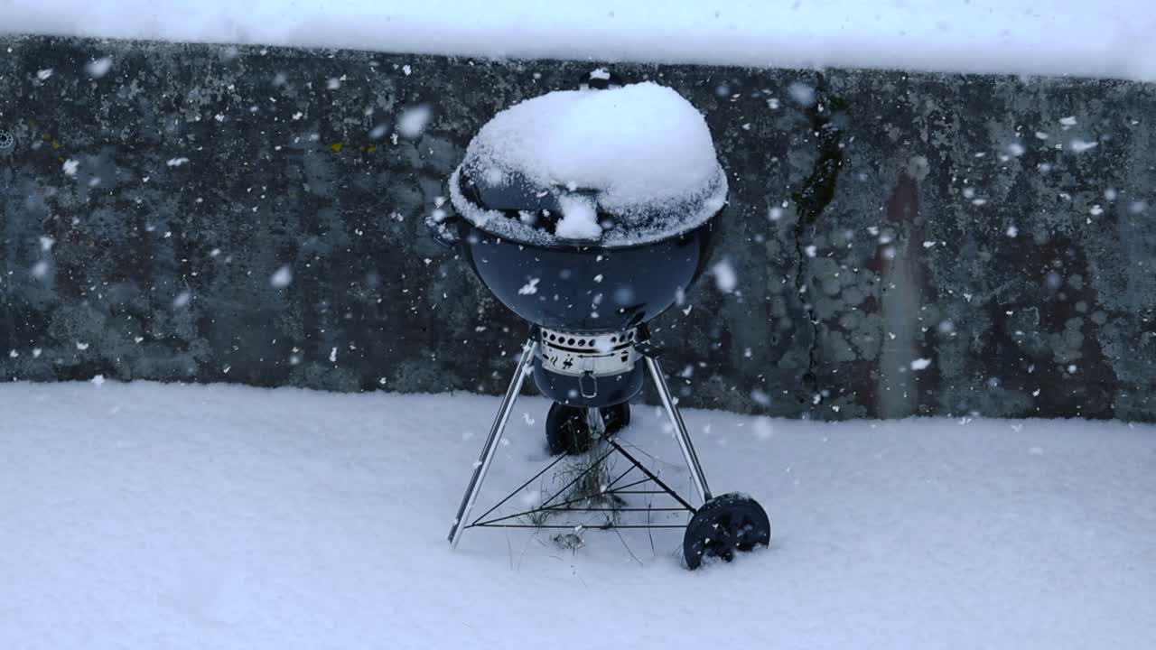 Slow motion of large snowflakes fall quietly covering the backyard ground with layer of snow. Outdoor closed round grill covered with snow, behind dark concrete wall texture. Black and white contrast