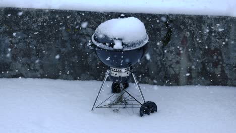 slow motion of large snowflakes fall quietly covering the backyard ground with layer of snow. outdoor closed round grill covered with snow, behind dark concrete wall texture. black and white contrast