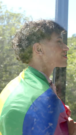 vertical video: smiling person wrapped in rainbow flag looking out window, embracing identity