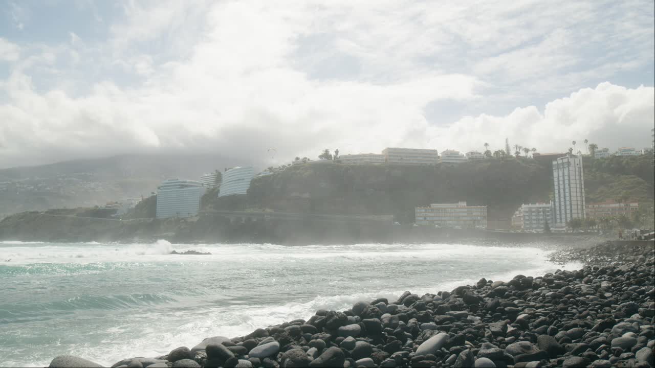 las olas del océano en cámara lenta aplastando la playa de guijarros, el parapente en tándem por encima de los resorts de hoteles, playa martiánez, puerto de la cruz, islas canarias en primavera