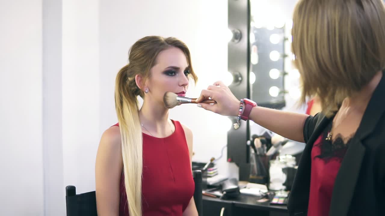 maquilladora usando un pincel grande para aplicar polvo facial y maquillaje de acabado para una joven rubia con vestido rojo. salón de belleza