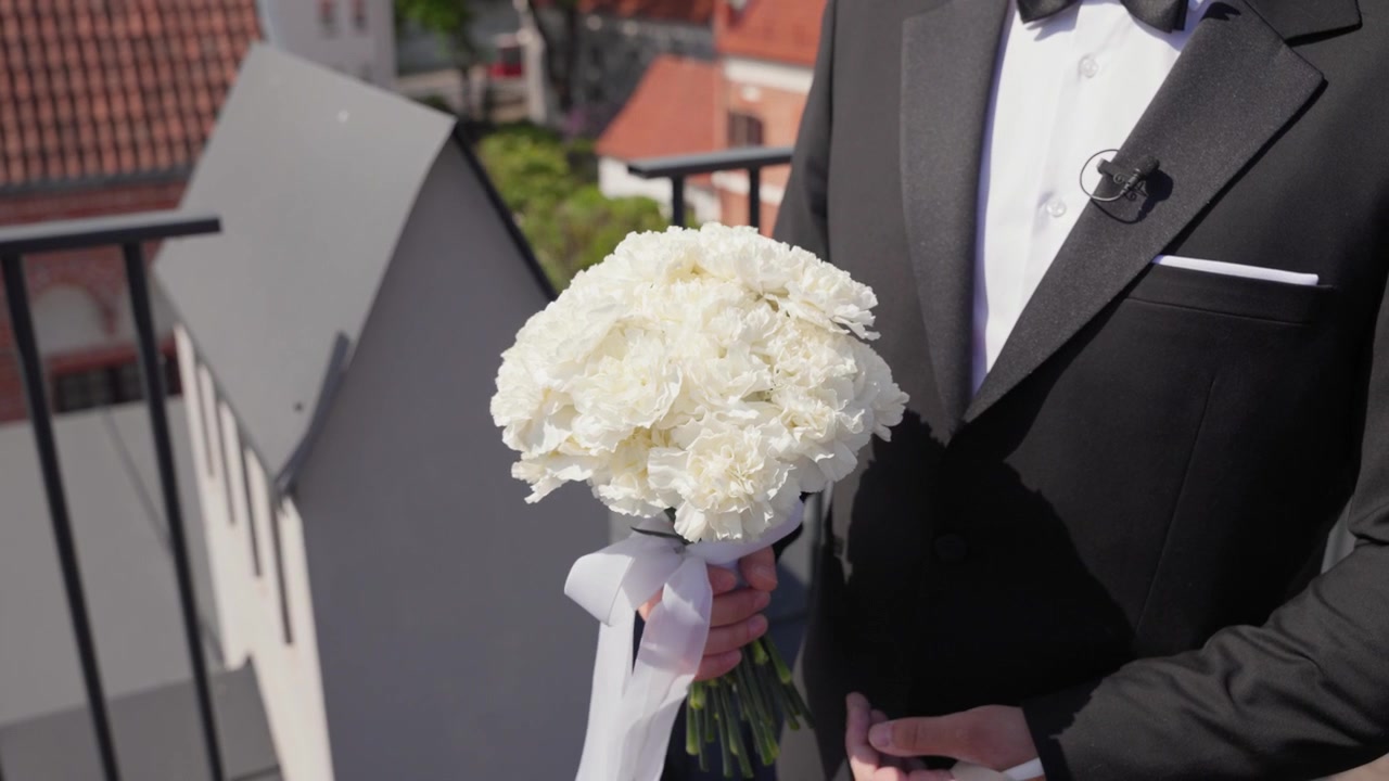 Uno sposo in un elegante smoking nero tiene un rigoglioso bouquet bianco con nastro di raso su un tetto illuminato dal sole, con lo skyline della città sullo sfondo. Luce naturale brillante, messa a fuoco nitida, ambientazione esterna romantica.
