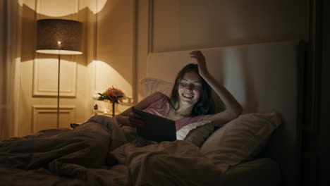 Cheerful woman watching tablet in bed. Happy female laughing movie in evening.