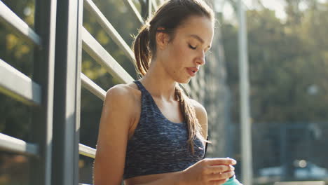 Mujer-Deportiva-Cansada-Parada-En-Una-Cancha-Al-Aire-Libre,-Descansando-Y-Bebiendo-Agua-Fría-Después-Del-Entrenamiento