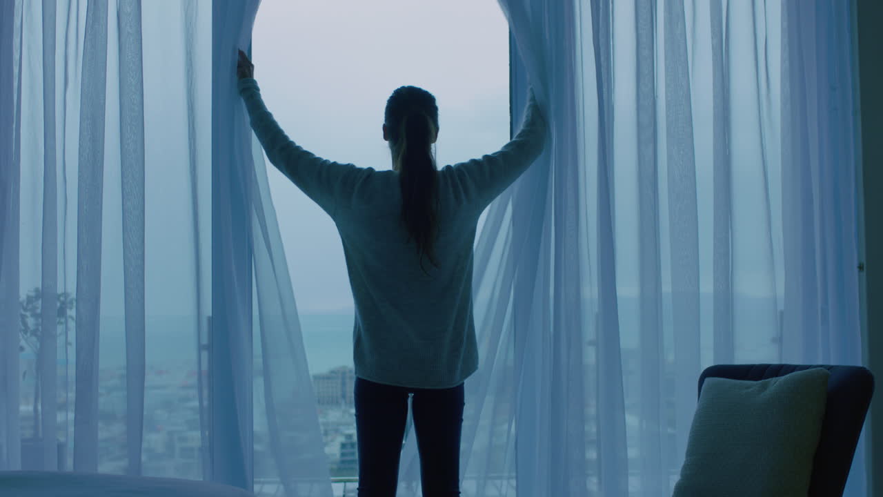 행복한 젊은 여성은 커튼을 열고 창문 밖을 바라보며 신선한 새로운 날을 준비하고 휴식을 취하고 도시의 전망을 즐기고 있습니다.