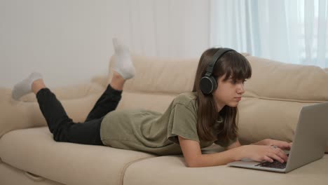 A-teen-girl-uses-a-laptop-while-lying-on-a-couch-in-headphones