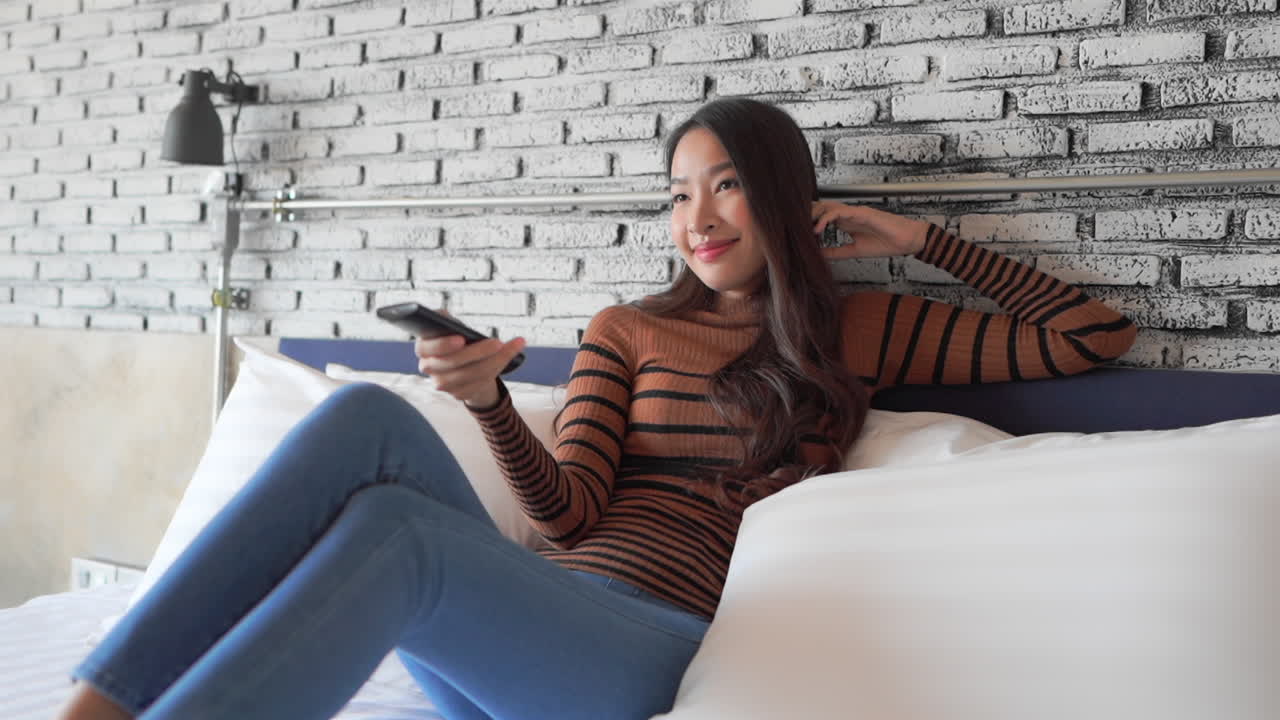 Pretty woman with a beautiful smile is changing TV channels holding remote control unit sitting on a white hotel bed