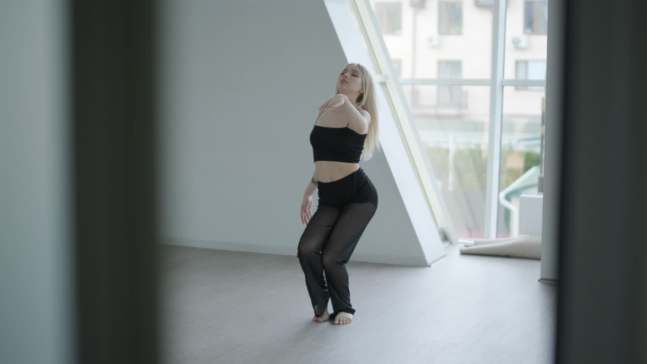 Trænet pige i sort tøj udfører moderne dans. Kvindelig danser viser flydende bevægelser, krop og hænder bølger i et hvidt rum. Moderne balletkoreografi. Ung kvinde bevæger sig smidigt
