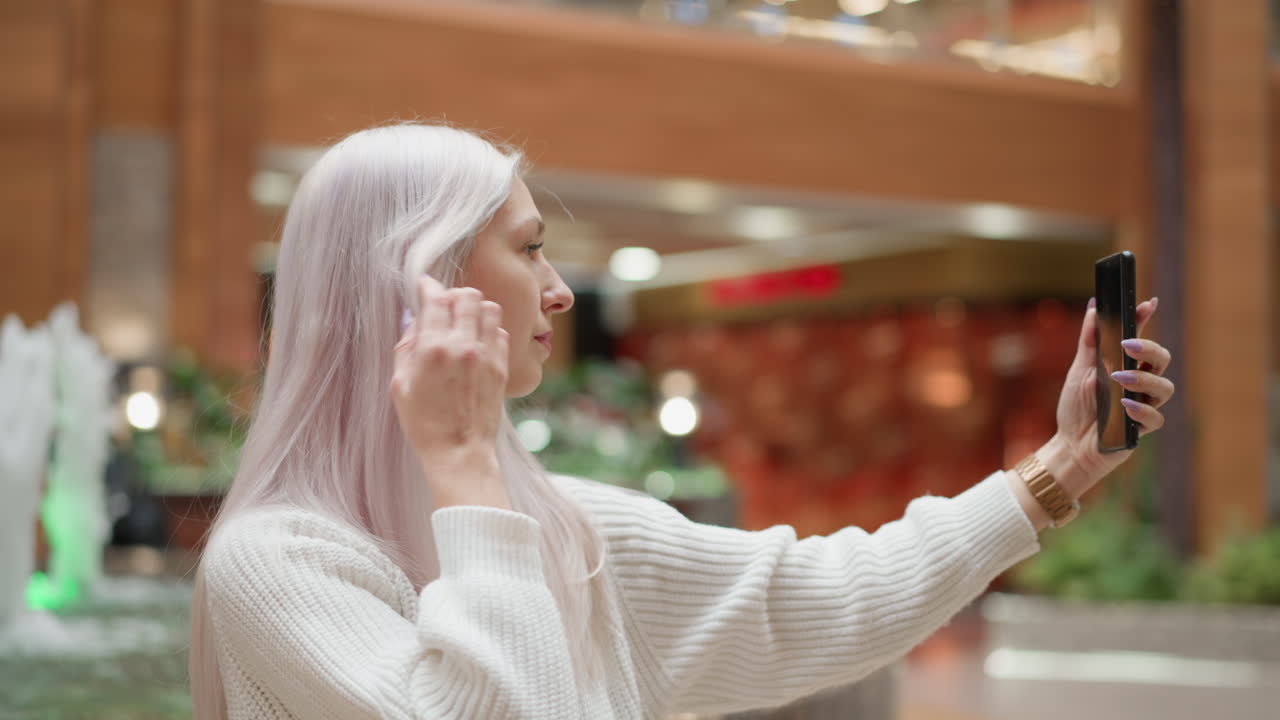Female shopper sits beside mall fountain on tiled bench, poses with mobile phone to capture water jets shimmering under lights, wearing sweater and jeans with polished nails and gold watch