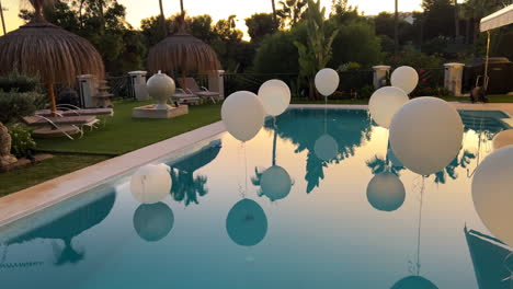 Hermosa-Fiesta-En-El-Jardín-De-La-Piscina-En-Un-Lugar-Tropical-Con-Globos-Blancos,-Sombrillas-Tiki-Y-Palmeras,-Reflejo-De-La-Puesta-De-Sol-En-El-Agua,-Celebración-Divertida,-Planificación-De-Eventos,-Tiro-De-4k