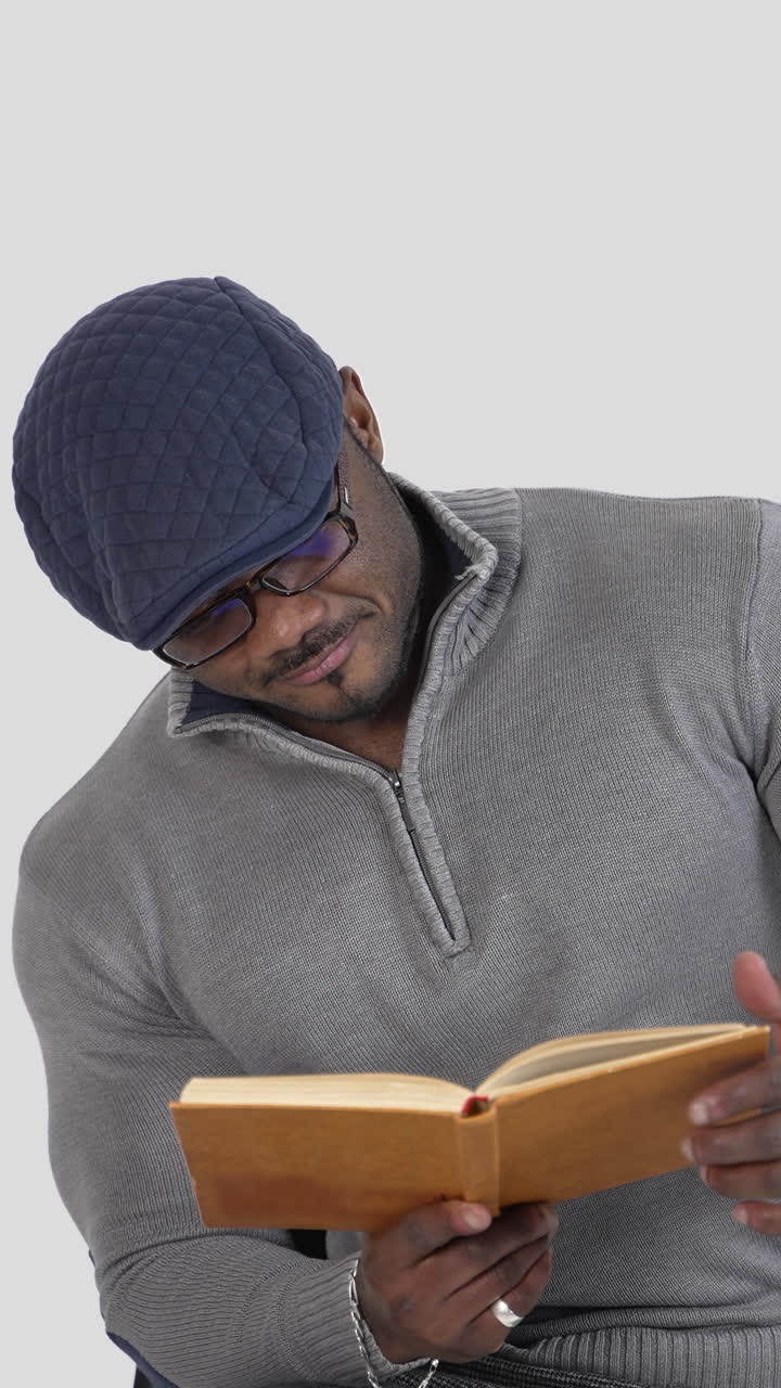 Serious african man reading book. Black skinned guy wearing glasses, cap and sweater sitting on chair with a book isolated on white. Vertical video