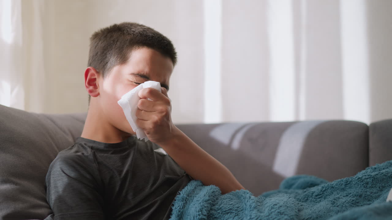 Boy lying down on couch, squeezing tissue on his nose to stop runny nose, surrounded by used tissues, feeling unwell, covered in blanket during flu recovery at home