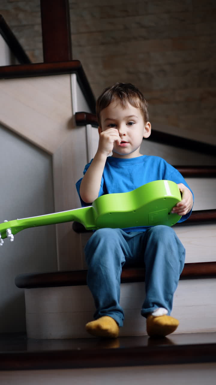 Calm Caucasian toddler boy sits on the stairs at home. Kid is holding a little light green guitar on his laps. Low angle view. Vertical video.