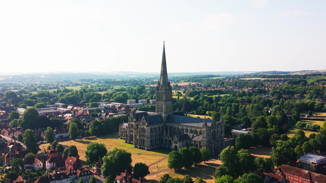 Ampia vista drone della Cattedrale di Salisbury e della sua guglia medievale, circondata da tetti storici, spazi verdi e la campagna del Wiltshire in una luminosa giornata estiva.
