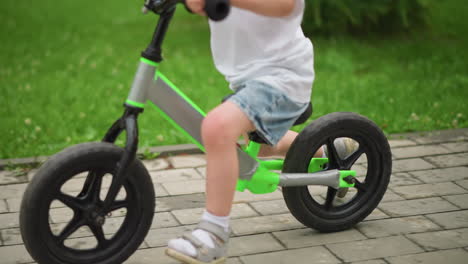 lower view of a young child riding a balance bike with his legs propelling on a paved walkway near lush greenery