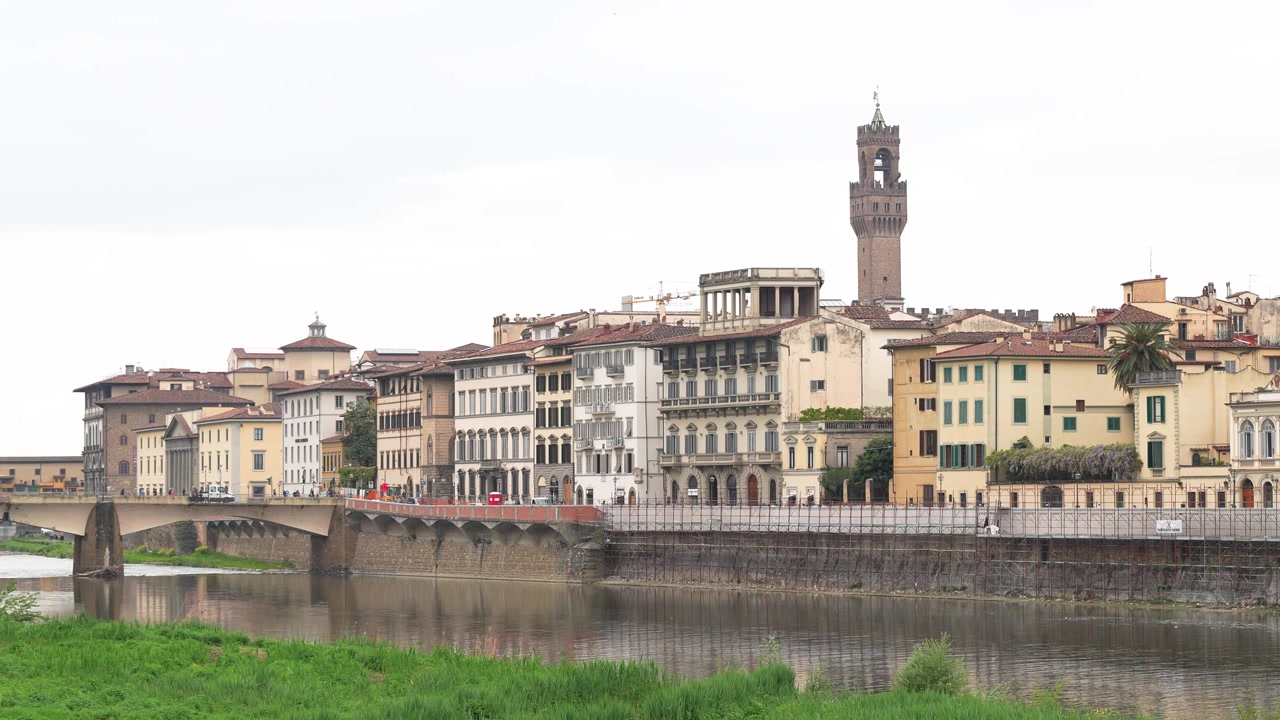 Ampia ripresa stabile del lungarno di Firenze che mostra colorati edifici rinascimentali, l'iconica torre di Palazzo Vecchio e le calme acque dell'Arno sotto una luce diurna soffusa, ideale per filmati di paesaggi urbani
