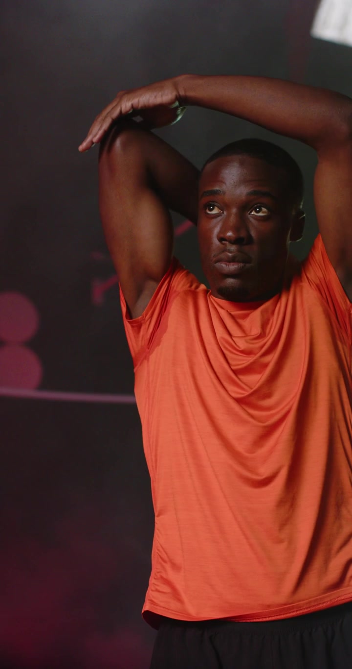 Vertical video: African American man warming, stretching chest and overhead in orange tee, studio