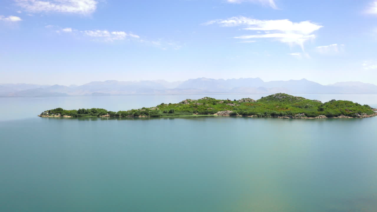 A small island in lake Skadar in Montenegro covered with lush vegetation and rocky outcrops, washed by clear blue waters on a sunny day, hazy mountains on the horizon, aerial zooming view 4K